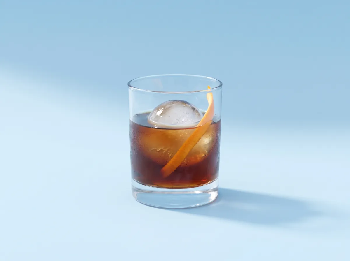 Cold brew in a short glass over a clear ice sphere with a twist of orange peel, on a sky-blue backdrop.